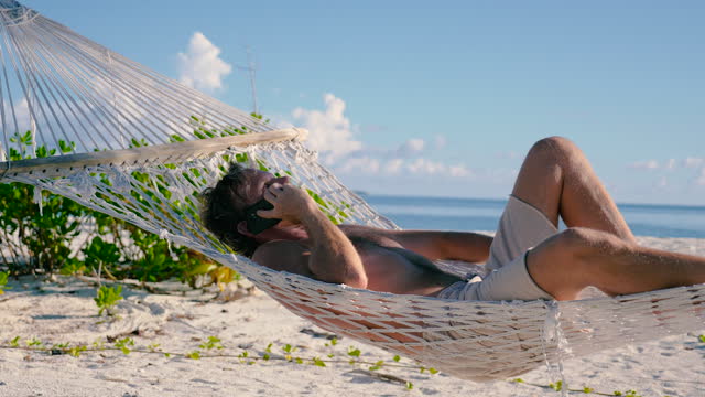 Man laying on a hammock between two palm trees on a beach taking a phone call