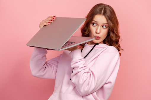 Portrait of curious curly haired teenage girl in hoodie peeping at laptop screen, shocked web content. Indoor studio shot isolated on pink background