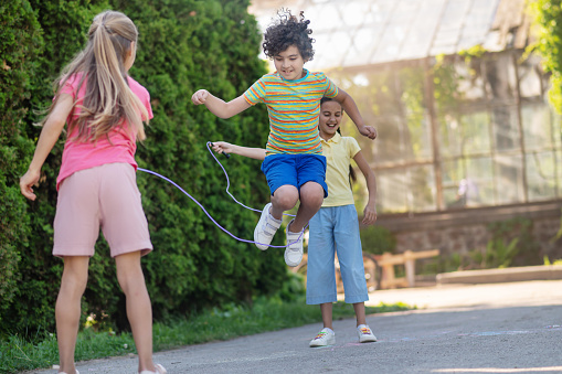 Jump rope. Energetic boy with dark curly hair in jump and two long-haired girls with skipping rope near green bushes