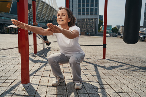 Smiling mature sporty woman doing powerful training near horizontal bars in open air on urban sports ground