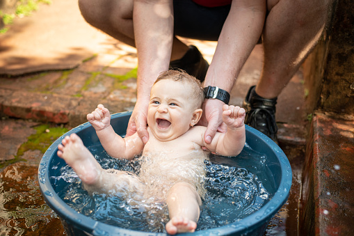 Father bathing baby in basin outside house during summer. Baby having so much fun.