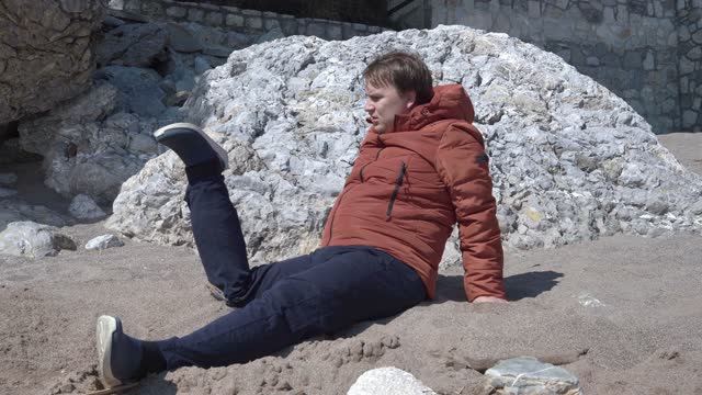 Plump man in orange jacket raises torso leaning on arms and looks at broken leg unusually bent near beach rocks on sunny spring day