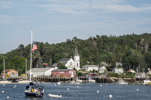 Boothbay, Maine, USA-July 12, 2021: Catholic church on shore of Boothbay Harbor, Maine. Lobster and fishing boats moored in the harbor.