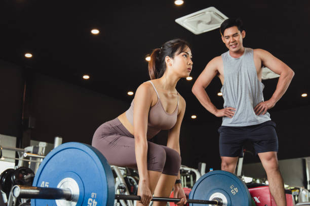 Asian women in sportswear preparing to lift barbells in fitness gym. Close up of young woman doing deadlift  workout with her handsome muscular personal trainer. Concept Exercise  for good shape stock photo