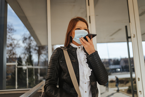 Portrait of one beautiful young business woman in white shirt with protective face mask standing in front of office building and talking on smart phone on sunny day