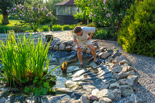 Young caucasian man cleans artificial garden pond bottom with landing net from mud, sludge and water plants. Spring and summer pond care work