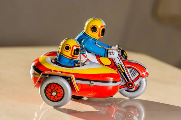 Wind-up old brass sidecar motorcycle toy showing two persons in it. Both persons wearing yellow helment. Pointing to the right.
Interior photo. Natural light. Toy shadow can be seen. No person