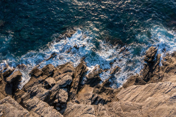 Deep blue waters swirl around rugged rocks, creating white foam as waves crash. Sunlight dances on the ocean, revealing a tranquil, natural beauty in a coastal paradise.