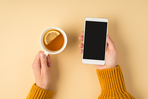First person top view photo of female hands in yellow sweater holding white cup of tea with lemon slice and smartphone on isolated light orange background with blank space