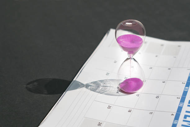 istock Hour glass with pink sand flowing and calendar on black tabletop background. 1330357923