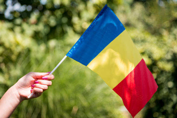 A female hand with painted nails holds the national flag of Romania, with lush green foliage defocused in the background.