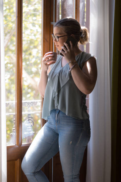 istock Young woman casually dressed in jeans by a window drinks coffee and talks on her phone. 1330362344