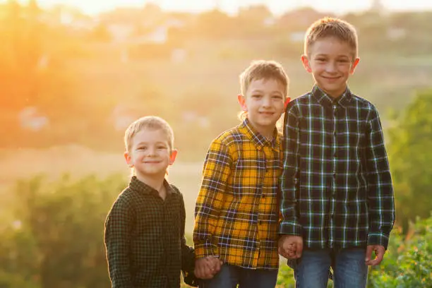 Portrait outdoor of three happy cheerful brothers at sunset. Guys in plaid shirts hold hands and smile while looking at camera. Friendly family concept.