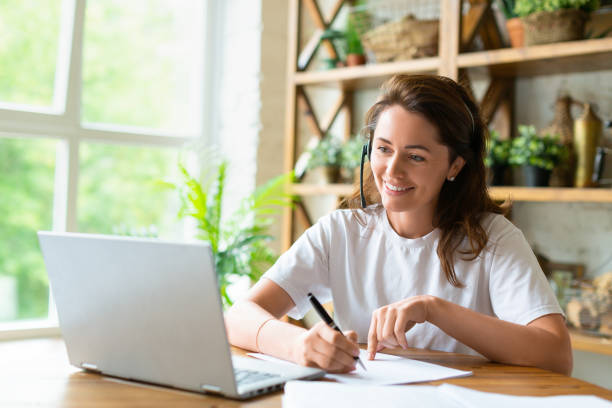 A smiling woman works with papers at home in front of a laptop monitor. She is checking papers during online video call stock photo
