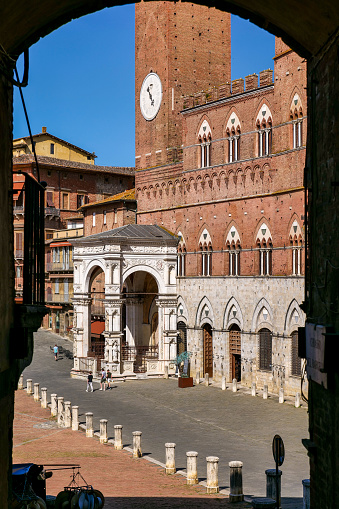 A stunning view of the Palazzo Pubblico in Piazza del Campo, in the historic heart of the medieval city of Siena, through one of the passages that allow access to the square through the buildings that surround it. Built starting in 1290 for the seat of the Council of Nine and the executive power of the Sienese city, the majestic Palazzo Pubblico is still the seat of the municipal government. Center of the ancient medieval life since 1169, the Piazza del Campo or simply Il Campo is one of the most beautiful and famous squares in the world for its particular shell shape. In this space every year the seventeen historic districts of Siena compete in the Palio, one of the oldest horse races in the world, during a representation of the medieval and historical celebrations of the city. Siena is one of the most beautiful Italian cities of art, in the heart of the Tuscan hills, visited for its immense artistic and historical heritage and for its famous popular traditions. Since 1995 the historic center of Siena has been declared a World Heritage Site by UNESCO. Image in high definition format.