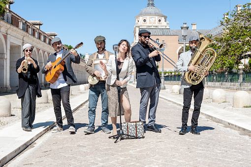 Jazz band playing in the street in broad daylight and dressed with jackets, caps and dresses; composed of 6 components, a singer, a clarinet, a tuba, a trombone, a guitar and a washboard. With the background of the gardens of Aranjuez, Madrid, Spain.