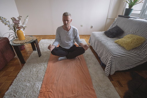 Adult man exercising at his living room on the floor. Meditating and resting in lotus yoga position.