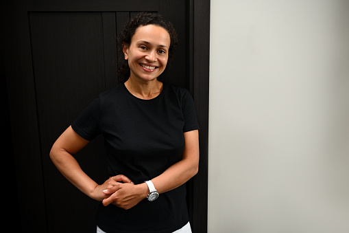 Middle aged mixed race African American woman, physiotherapist at a spa wellness center, wearing a black t-shirt, smiling cutely at the camera, standing in front of the door of her massage parlor