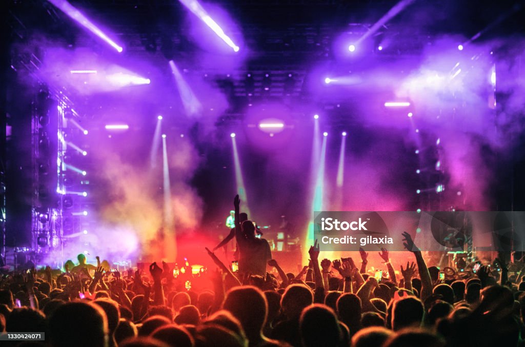 Large group of people at a concert party. Rear view of large group of people enjoying a concert performance. There are many hands applauding and taping the show. Blue and red spot lights firing from the stage.

Each individual inspected for recognizability. Music Festival Stock Photo