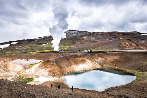 People walking hiking on Viti Crater of Krafla caldera geothermal formation with small lake water and mountains snow during summer and geyser hot spring steam