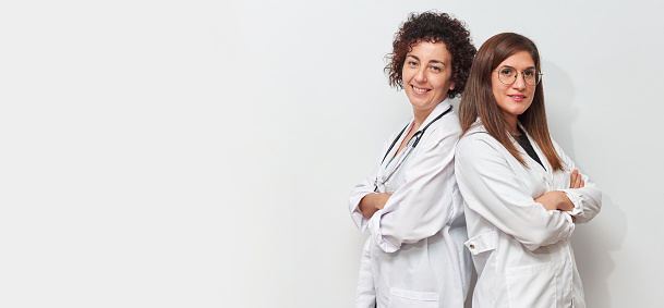 Portrait of two Caucasian female doctors back to back looking at camera dressed in lab coats