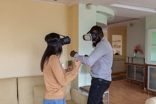 Excited Young Multi-Ethnic Couple is Having Fun with VR Glasses. Young Afro-American Man and his Asian Girlfriend are Experiencing Video Games on Virtual Reality Simulator.