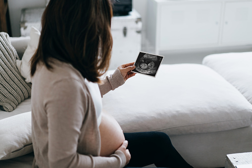 A loving young Asian pregnant woman relaxing on sofa at home, looking at the ultrasound scan photo of her baby while touching her belly. Mother-to-be. Expecting a new life concept