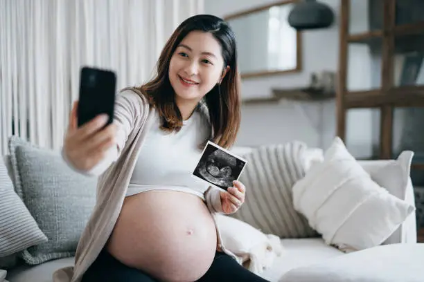 Joyful young Asian pregnant woman taking a selfie with smartphone, holding ultrasound scan photo on hand. Mother-to-be. Expecting a new life. Heathy pregnancy lifestyle