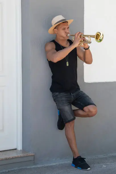 Photo of Latino man with a hat leaning on a wall playing the trumpet: Selective focus. Music concept.