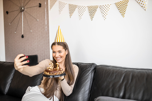 Beautiful woman having video call while celebrating her 21's birthday with chocolate cake at home