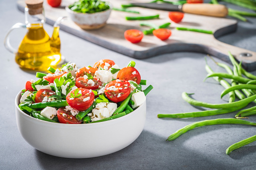 Healthy vegetarian salad bowl with cherry tomatoes, green beans, feta cheese and parsley with olive oil, cutting board and knife, Mediterranean diet