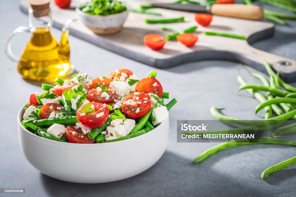 Healthy salad bowl with cherry tomatoes, green beans, feta cheese and parsley Mediterranean diet Healthy vegetarian salad bowl with cherry tomatoes, green beans, feta cheese and parsley with olive oil, cutting board and knife, Mediterranean diet Salad Stock Photo