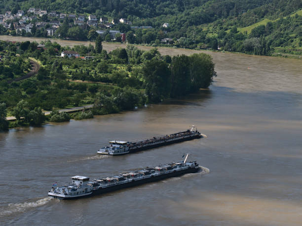 Boppard, Germany - 07-18-2021: Two inland cargo ships sailing alongside on Rhive river, an important waterway, on sunny day with town Boppard in background and green trees in summer.