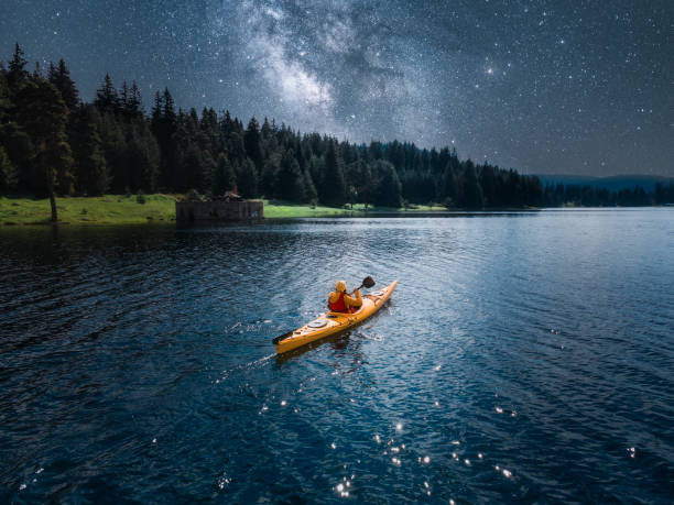 Woman kayaking in mountain lake under Milky way. Aerial view with starry sky. Paddling and ecotourism. Drone point of view directly above woman with yellow kayak rowing and connecting with nature at night. Exercise and body care. kayaking stock pictures, royalty-free photos & images