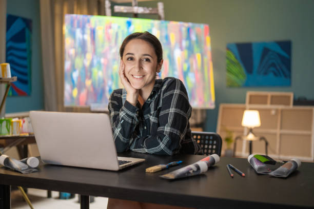portret uśmiechniętej brunetki ubranej w koszulę w kratkę siedzącej przy biurku przed laptopem w studiu sztuki i podpierającej się ręką, na łokciu pod brodą, patrzącej w kamerę - propping zdjęcia i obrazy z banku zdjęć