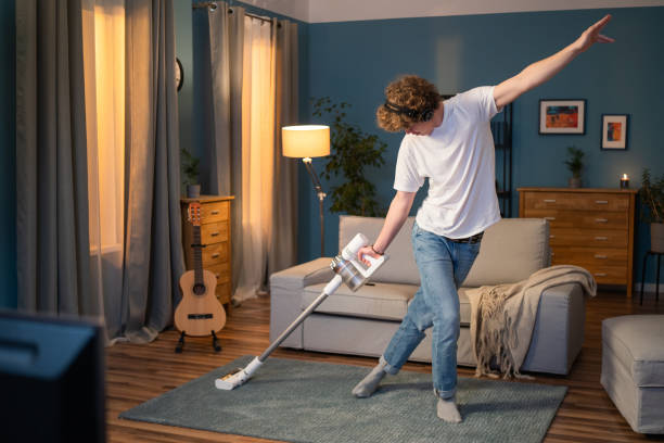 한 소년이 저녁에 거실을 청소하고 있습니다. 남자는 방에 카펫을 진공 청소기, 무선 헤드폰에 음악을 듣고 진공 청소기와 함께 춤을 재미있다 - new housing audio 뉴스 사진 이미지