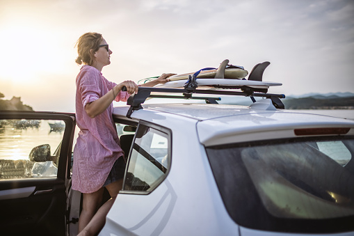 Photo of young woman packing her surfboards on the top of the car, after long day at the beach.