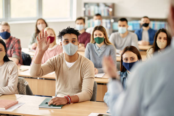 Black student with face mask raising hand to ask a question during lecture at college classroom. African American student raising his hand to answer a question while attending lecture in the classroom during COVID-19 pandemic. hand raised classroom student high school student stock pictures, royalty-free photos & images