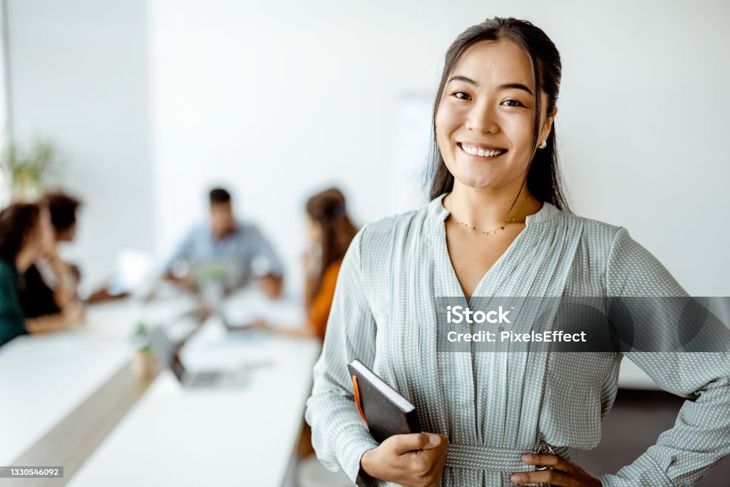 Doing business with a smile Confident young Asian businesswoman standing smiling at the camera in a boardroom with colleagues in the background Employee Stock Photo