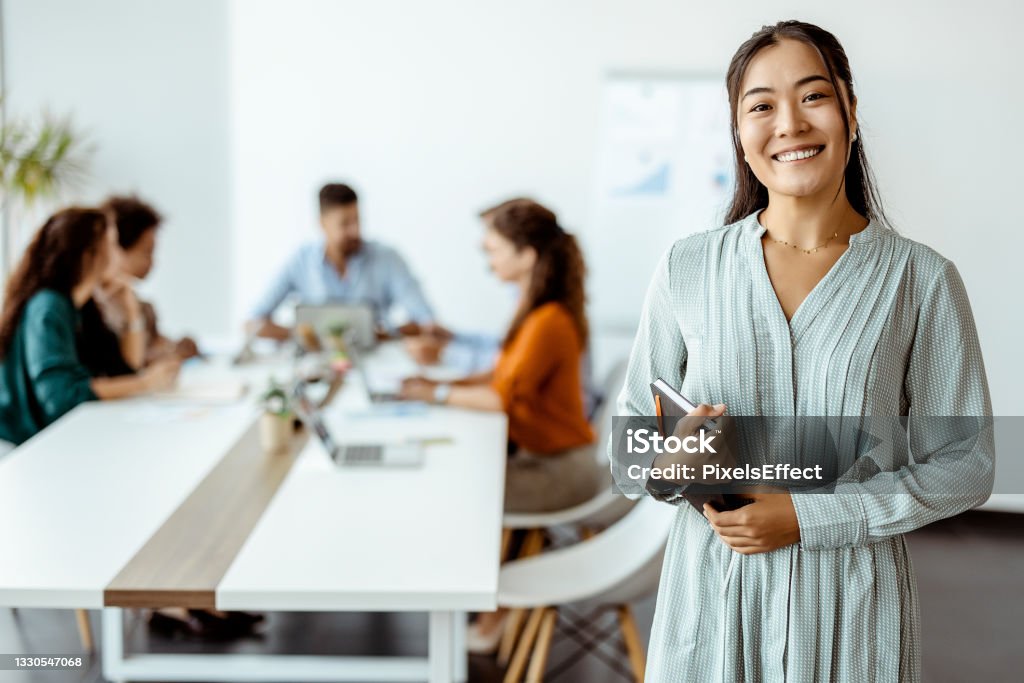 Doing business with a smile Confident young Asian businesswoman standing smiling at the camera in a boardroom with colleagues in the background Office Stock Photo