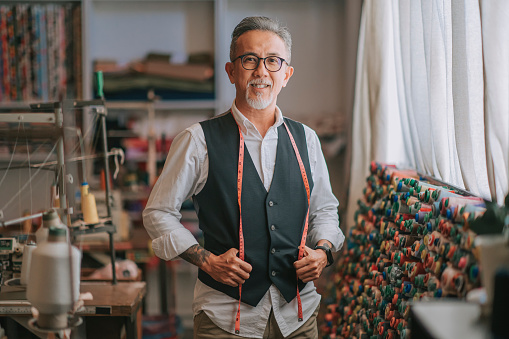 asian chinese senior tailor man with facial hair looking at camera smiling in atelier studio