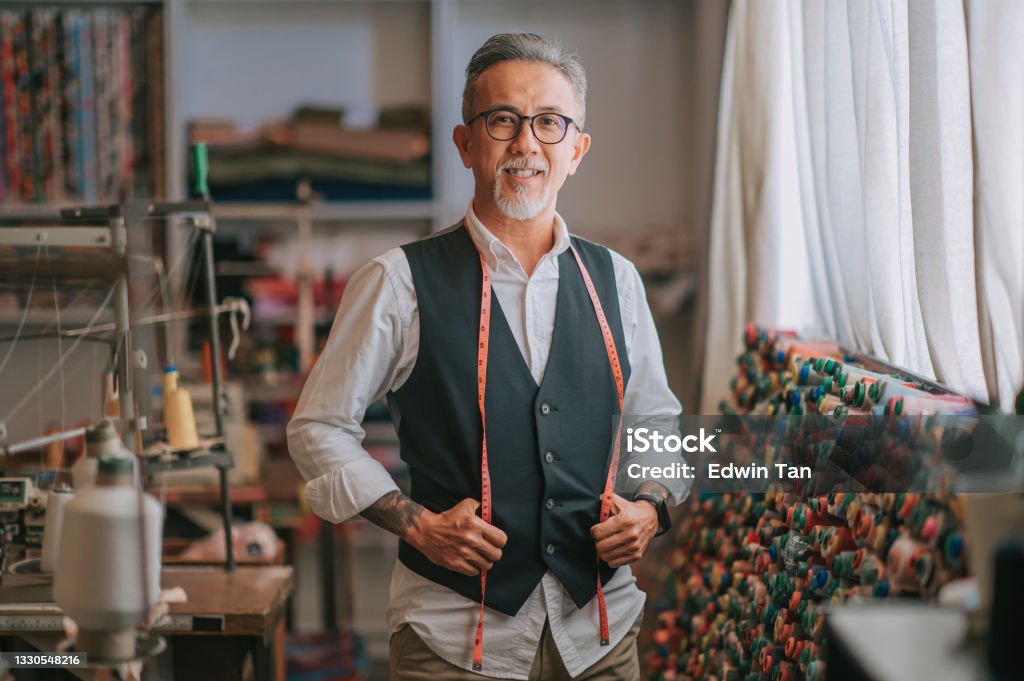 asian chinese senior tailor man with facial hair looking at camera smiling in atelier studio Owner Stock Photo