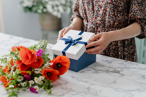 Cropped shot of woman wearing dress opening gift box with blue ribbon and bow, taking white top off, peeking inside, marble table with colorful flower bouquet, blurred background