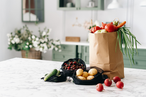 Various fresh vegetables in paper grocery and black mesh bags on kitchen island with marble top, healthy food full of nutrients, vintage furniture with flowers, blurred background
