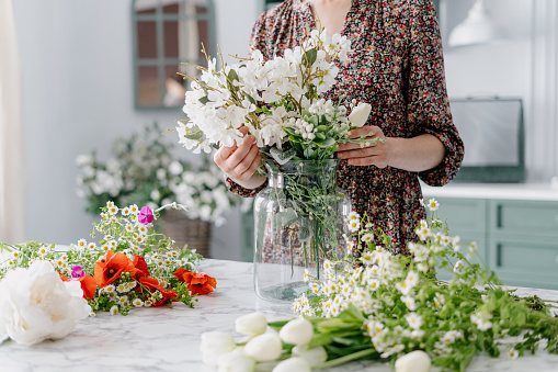 Marble kitchen island with variety of colorful flowers such as tulips, poppies and chamomiles laying around, woman in dress putting them in big glass jar, green counter in blurred background