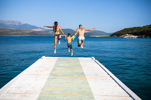 A young mother and father are holding their small sun by the hand and jumping off a pier into the sea