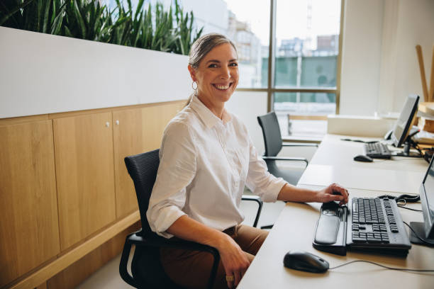 friendly receptionist working in office - senior adult happiness computer looking at camera imagens e fotografias de stock