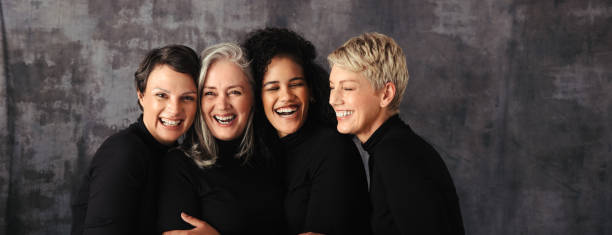 Smiling women of different ages standing in a studio Smiling women of different ages standing together in studio. Group of four confident women embracing their natural and ageing bodies against a studio background. mixed age range stock pictures, royalty-free photos & images