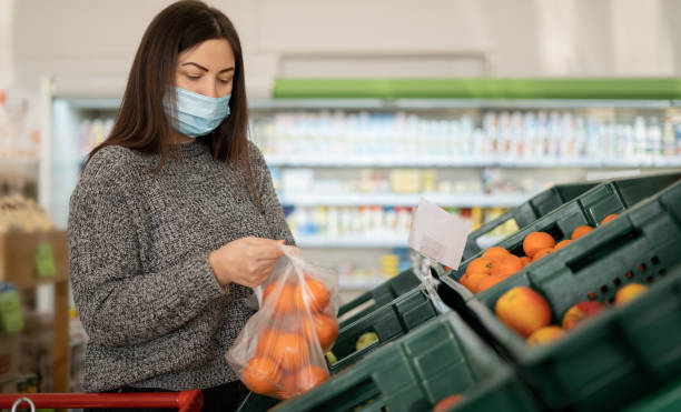 a young girl wearing a protective medical mask is shopping at the supermarket and putting citrus fruits in a bag in the fruits and vegetables department. - retail occupation flash imagens e fotografias de stock