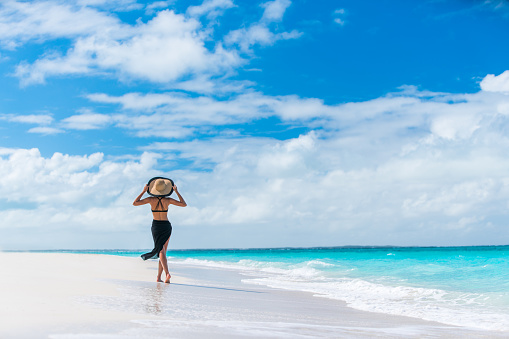 Luxury travel woman in black beachwear sarong walking taking a stroll on perfect white sand Caribbean beach. Girl tourist on summer holiday holding sun hat at vacation resort. Tropical landscape.